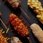 Korean Corn Dog varieties with different toppings like ketchup, mustard, sugar, and potato fries on a black background.