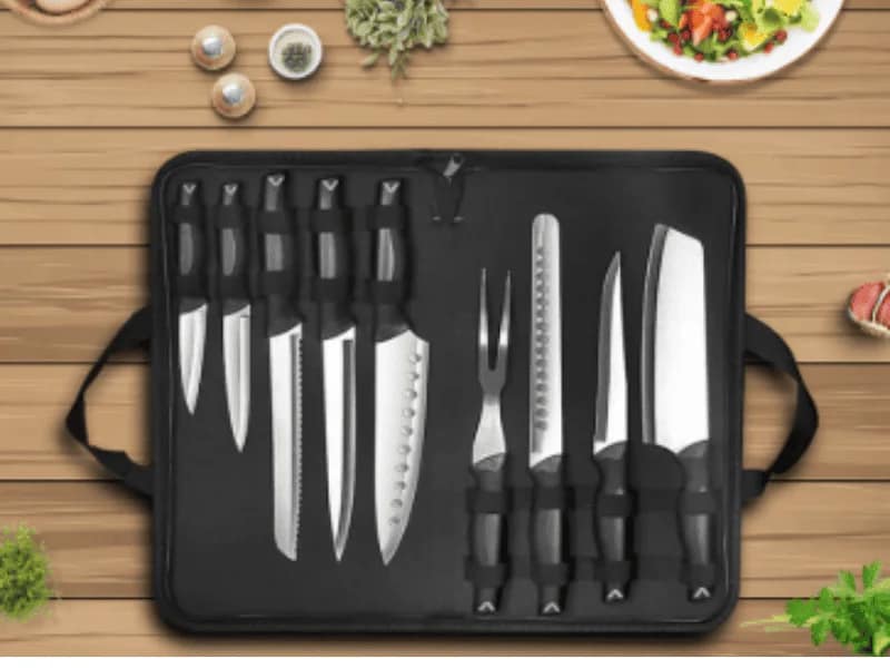 Professional Hell kitchen knife set in a black carrying case placed on a wooden table with herbs and salad around.