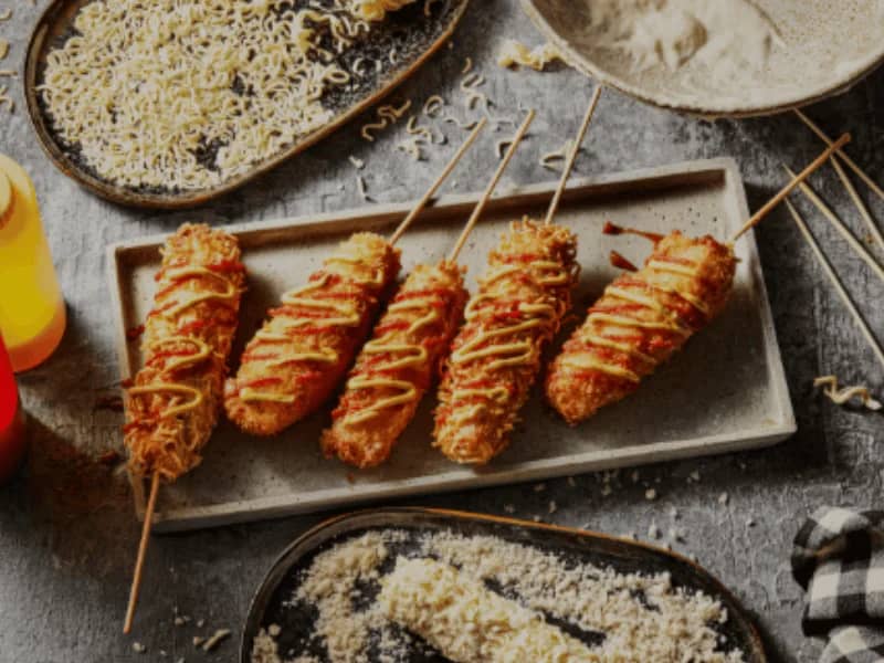 Korean Corn Dog served on a tray with ketchup and mustard, surrounded by cheese, flour, and ramen crumbs.