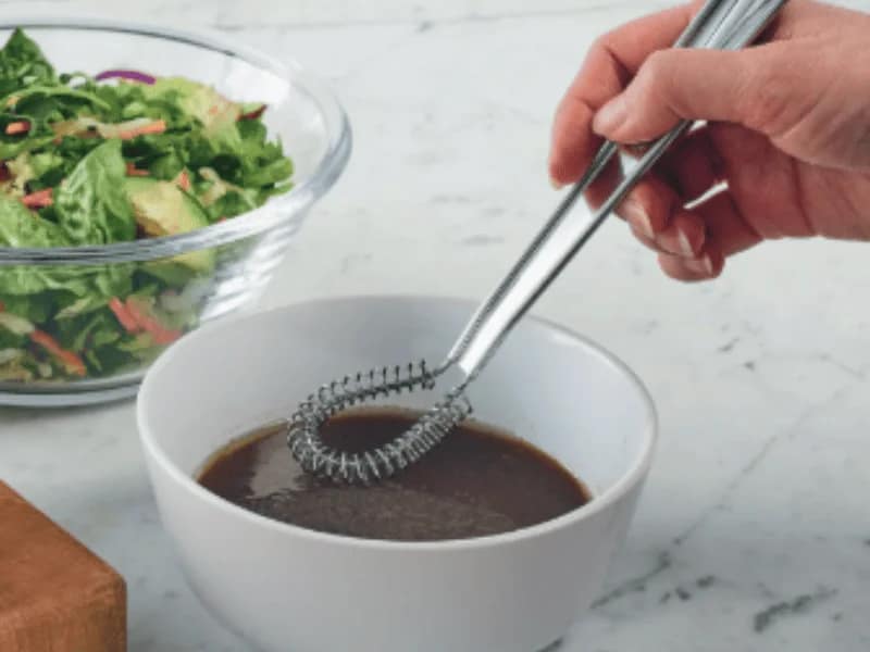 Whisks – A hand using a stainless steel whisk to stir salad dressing in a white bowl, with a glass bowl of salad nearby.