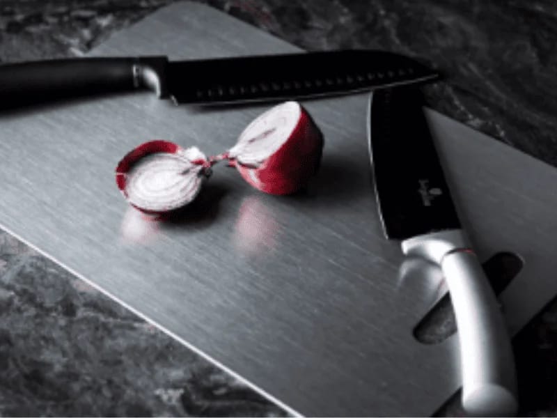 Titanium Cutting Board with two kitchen knives and a sliced red onion on a dark countertop.
