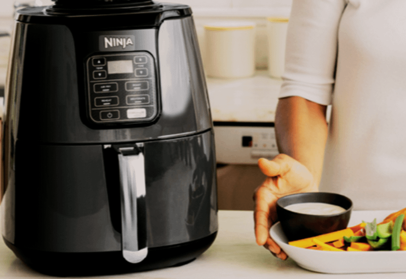 Air Fryers by Ninja with digital controls on a kitchen counter beside a person holding a plate of fresh vegetables and dip.